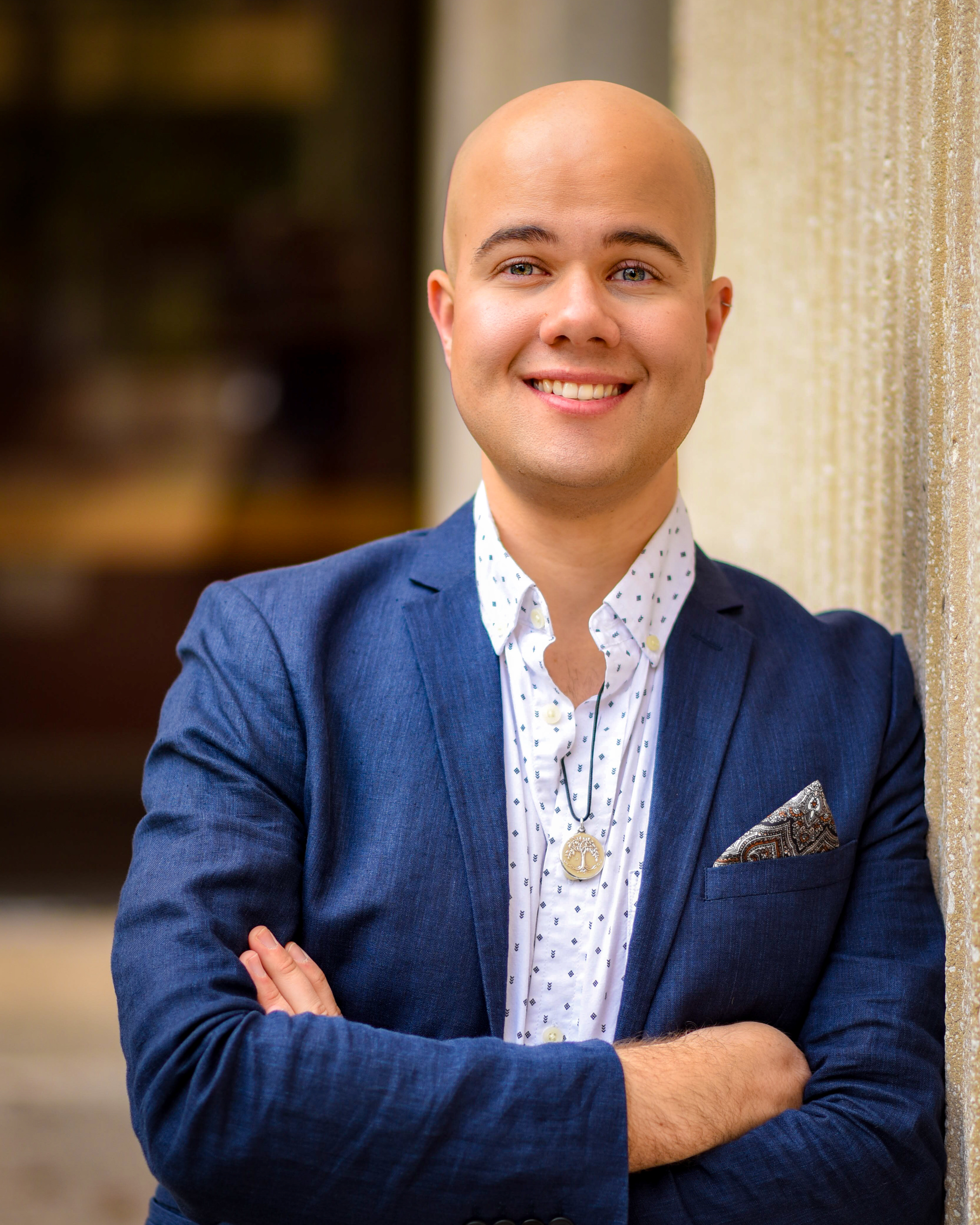 A bald man with a blue suit jacket and white dress shirt leans up against a grey wall. Their arms are folded across their chest and they are smiling into the camera.